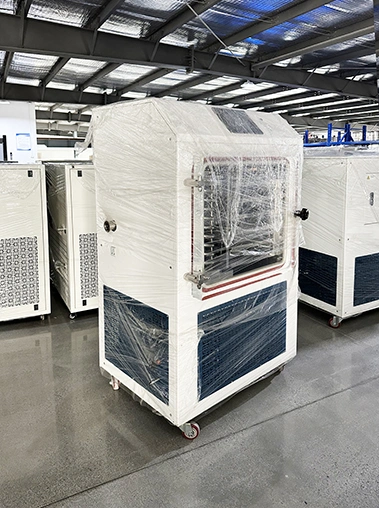 Left perspective view of a white pilot vacuum freeze dryer wrapped in protective plastic film inside a factory workshop.